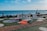 A romantic seaside setup features large letters spelling 'MARRY ME' in front of an ocean backdrop. Decorative elements include a picnic setting with a red blanket and cushions, surrounded by white circular stone patterns on the grass. There are also small candle holders placed around the blanket, adding to the intimate atmosphere. Two angel statues stand on either side, and there are ships visible in the distance on the ocean.