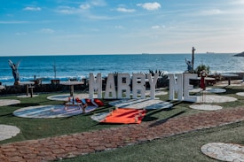 A romantic seaside setup features large letters spelling 'MARRY ME' in front of an ocean backdrop. Decorative elements include a picnic setting with a red blanket and cushions, surrounded by white circular stone patterns on the grass. There are also small candle holders placed around the blanket, adding to the intimate atmosphere. Two angel statues stand on either side, and there are ships visible in the distance on the ocean.