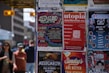 A collection of colorful posters is displayed on a wall, advertising various events and products. The posters include promotions for music festivals, a mushroom company, an anime festival, and a concert tour. People are casually walking by in the background, suggesting an urban setting.
