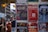 A collection of colorful posters is displayed on a wall, advertising various events and products. The posters include promotions for music festivals, a mushroom company, an anime festival, and a concert tour. People are casually walking by in the background, suggesting an urban setting.