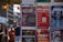 A collection of colorful posters is displayed on a wall, advertising various events and products. The posters include promotions for music festivals, a mushroom company, an anime festival, and a concert tour. People are casually walking by in the background, suggesting an urban setting.