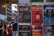 A collection of colorful posters is displayed on a wall, advertising various events and products. The posters include promotions for music festivals, a mushroom company, an anime festival, and a concert tour. People are casually walking by in the background, suggesting an urban setting.