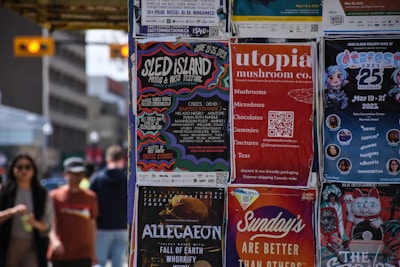 A collection of colorful posters is displayed on a wall, advertising various events and products. The posters include promotions for music festivals, a mushroom company, an anime festival, and a concert tour. People are casually walking by in the background, suggesting an urban setting.