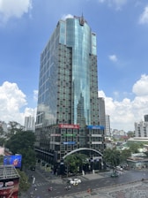 A tall modern office building with a glass facade, featuring various company logos on the exterior. It's situated in a bustling urban area with trees and smaller buildings nearby. The sky is partly cloudy.