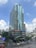 A tall modern office building with a glass facade, featuring various company logos on the exterior. It's situated in a bustling urban area with trees and smaller buildings nearby. The sky is partly cloudy.