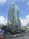 A tall modern office building with a glass facade, featuring various company logos on the exterior. It's situated in a bustling urban area with trees and smaller buildings nearby. The sky is partly cloudy.