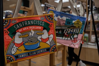 Tote bags featuring whimsical illustrations hang on display. The first bag showcases two cartoon birds enjoying a bowl of steaming clam chowder with the text 'San Francisco Bay Area Fresh Catch, Famous Clam Chowder'. Another bag has an illustration of a trolley in front of colorful buildings, capturing an iconic San Francisco scene.