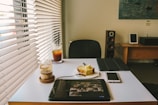 A bright room with a modern interior features a table displaying a tablet, a smartphone, a plate with a slice of cake, a glass of layered coffee, and iced tea. Natural light streams in through the blinds on the window. In the background, there's a loudspeaker, a small framed picture, and a wooden desk with another framed picture and electronic device.