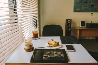 A bright room with a modern interior features a table displaying a tablet, a smartphone, a plate with a slice of cake, a glass of layered coffee, and iced tea. Natural light streams in through the blinds on the window. In the background, there's a loudspeaker, a small framed picture, and a wooden desk with another framed picture and electronic device.