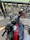 A motorcycle handlebar and speedometer with a decorative cloth tied to it. The fuel tank has a shiny black surface with a red stripe. A wrought iron railing and stone pavement are visible in the background along with a body of water.