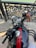 A motorcycle handlebar and speedometer with a decorative cloth tied to it. The fuel tank has a shiny black surface with a red stripe. A wrought iron railing and stone pavement are visible in the background along with a body of water.