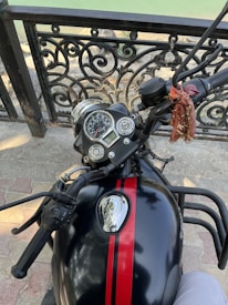 A motorcycle handlebar and speedometer with a decorative cloth tied to it. The fuel tank has a shiny black surface with a red stripe. A wrought iron railing and stone pavement are visible in the background along with a body of water.