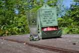 A clear glass with a logo from the Point Defiance Zoo & Aquarium sits on a weathered wooden table beside a vintage green metal holder featuring text for a grain and feed elevator company. The background is filled with lush green foliage under a clear blue sky.