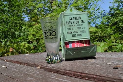 A clear glass with a logo from the Point Defiance Zoo & Aquarium sits on a weathered wooden table beside a vintage green metal holder featuring text for a grain and feed elevator company. The background is filled with lush green foliage under a clear blue sky.