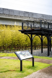 An elevated metal platform with a railing stands in front of a dense bamboo grove. A concrete structure, possibly a bridge or railway, extends above. A signboard with informational panels is positioned on the grassy area below.