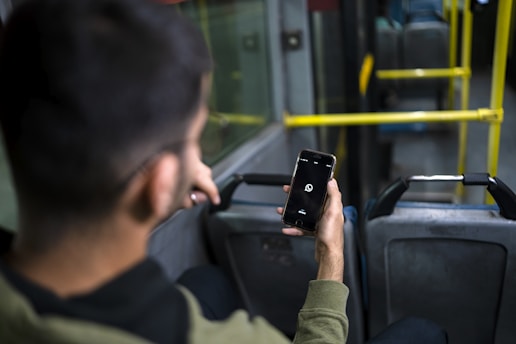 A person is seated on a bus holding a smartphone with a messaging app open on the screen. The interior of the bus is visible, featuring gray and yellow seating and poles for stability.