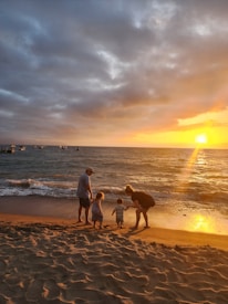 A family of four, including two adults and two children, stands on a sandy beach by the ocean at sunset. The golden sunlight illuminates the sandy shore and casts long shadows. Several boats are visible on the horizon, framed by a dramatic sky with dark clouds and a vibrant sunset.