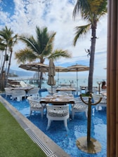 A picturesque outdoor dining area with tables set over a shallow pool, surrounded by elegant white chairs. Large beige umbrellas provide shade, while tall palm trees add a tropical ambiance. The background features a clear view of the ocean and a blue sky with scattered clouds.
