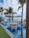A picturesque outdoor dining area with tables set over a shallow pool, surrounded by elegant white chairs. Large beige umbrellas provide shade, while tall palm trees add a tropical ambiance. The background features a clear view of the ocean and a blue sky with scattered clouds.