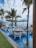 A picturesque outdoor dining area with tables set over a shallow pool, surrounded by elegant white chairs. Large beige umbrellas provide shade, while tall palm trees add a tropical ambiance. The background features a clear view of the ocean and a blue sky with scattered clouds.