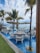 A picturesque outdoor dining area with tables set over a shallow pool, surrounded by elegant white chairs. Large beige umbrellas provide shade, while tall palm trees add a tropical ambiance. The background features a clear view of the ocean and a blue sky with scattered clouds.