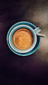 A top-down view of a blue coffee cup filled with espresso, placed on a dark surface. The coffee has a rich crema on top, indicating a well-made brew.