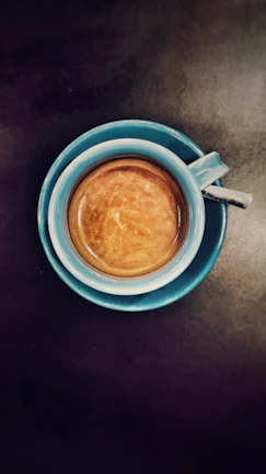 A top-down view of a blue coffee cup filled with espresso, placed on a dark surface. The coffee has a rich crema on top, indicating a well-made brew.