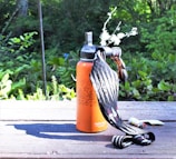 A bright orange water bottle sits on a wooden surface surrounded by lush green vegetation in an outdoor setting. A patterned cloth and small personal items, including a lighter, are also present on the table.