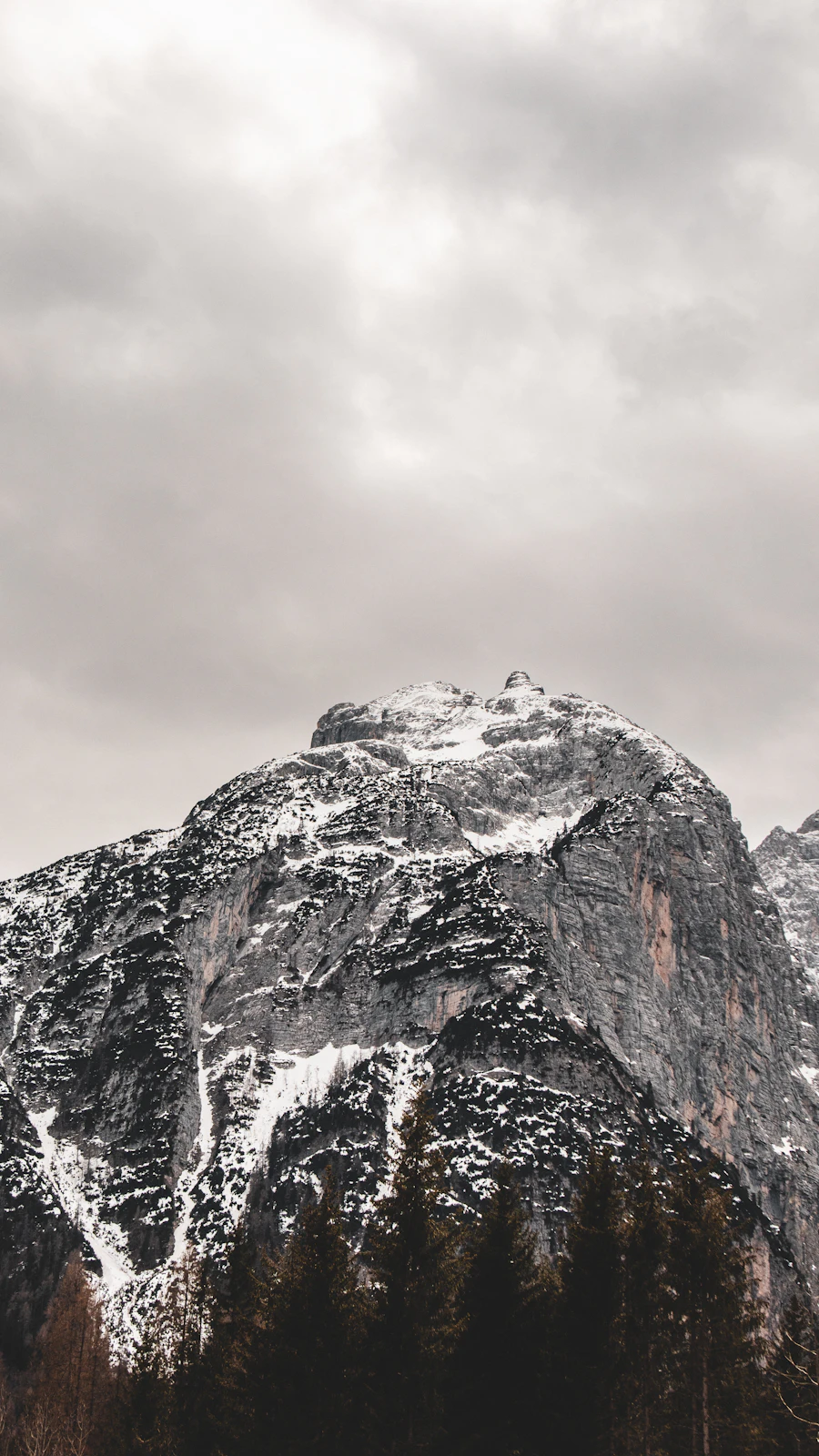Snow-dusted grey Dolomite rock face rising above dark pine forest under heavy cloud