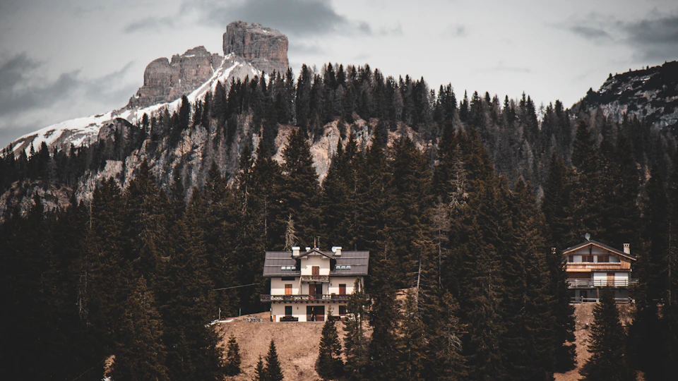 The image depicts a serene mountain landscape with a small wooden cabin nestled among tall trees on a hillside, surrounded by snow-capped mountains and a cloudy sky. The cabin has a white roof and brown walls, blending harmoniously with the natural surroundings. In the distance, more trees can be seen, adding depth to the scene.