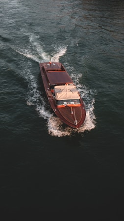 A wooden speedboat navigates through calm, dark waters, creating gentle waves. The boat is sleek in design with a classic wooden finish and a beige canopy.