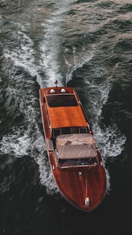 A sleek, wooden speedboat glides swiftly across a body of water, leaving a trail of frothy waves in its wake. The surface of the water is deep green, contrasting with the rich, polished texture of the boat's wooden deck. A small flag is visible at the stern of the boat.
