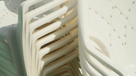 A stack of white plastic chairs arranged in a neat pile. The chairs have a simple, modern design with curved backs and small drainage holes.