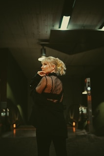 A person with blonde hair and an intricate hairstyle stands in an urban setting, wearing a black outfit with a translucent, dotted back. The background features industrial lighting and a metallic structure, creating a moody and sophisticated atmosphere.
