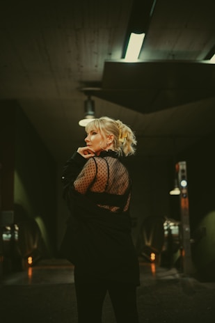 A person with blonde hair and an intricate hairstyle stands in an urban setting, wearing a black outfit with a translucent, dotted back. The background features industrial lighting and a metallic structure, creating a moody and sophisticated atmosphere.