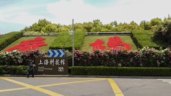 A landscaped display with large, red floral designs integrated into green hedges, set against a backdrop of trees. A pedestrian walks past a sign that directs visitors to the Shanghai Science & Technology Museum's gate number 3.