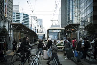 A busy city street featuring a tram station with people crossing the tracks. The background shows tall modern buildings and commuters on bicycles. Some individuals carry luggage, suggesting a bustling urban environment.
