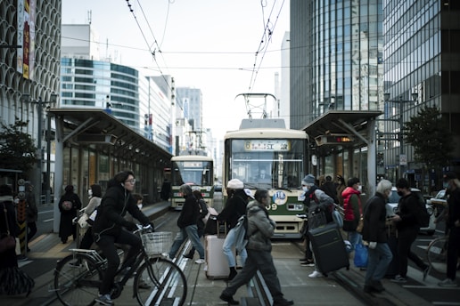 A busy city street featuring a tram station with people crossing the tracks. The background shows tall modern buildings and commuters on bicycles. Some individuals carry luggage, suggesting a bustling urban environment.