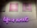 A wall with two framed posters and a neon sign. The posters have humorous text about friends, waffles, and work, and are on a pink background. Below them is a neon sign in purple light that reads 'life is sweet'. The wall has a white brick texture.