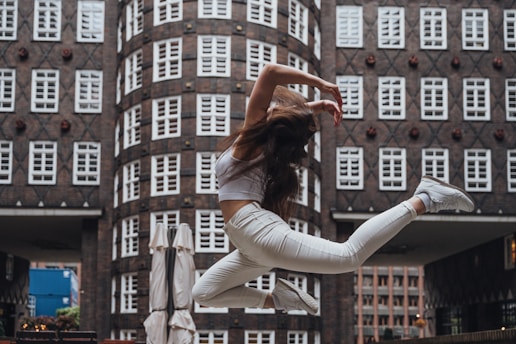 A person wearing a white outfit leaps gracefully in mid-air against a backdrop of an urban building with numerous windows. The motion captures a sense of freedom and dynamism, with the person's hair flowing and limbs extended in an elegant pose.
