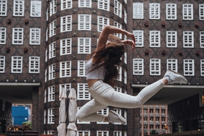A person wearing a white outfit leaps gracefully in mid-air against a backdrop of an urban building with numerous windows. The motion captures a sense of freedom and dynamism, with the person's hair flowing and limbs extended in an elegant pose.