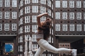 A person is captured mid-air in a dynamic dance pose against a backdrop of an urban architectural structure with symmetrical windows and brickwork.