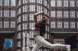 A person is captured mid-air in a dynamic dance pose against a backdrop of an urban architectural structure with symmetrical windows and brickwork.