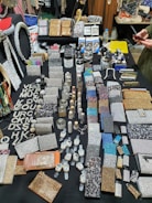 An assortment of sparkling and glittery accessories is displayed on a black table. The items include wallets, vanity cases, compact mirrors, and wristwatches, among others. Many pieces are embellished with rhinestones, sequins, and dazzling patterns. A variety of colors such as silver, gold, and multicolored designs are present, and the overall display is organized in a visually appealing manner. Some text made from shiny material is seen on the left side.