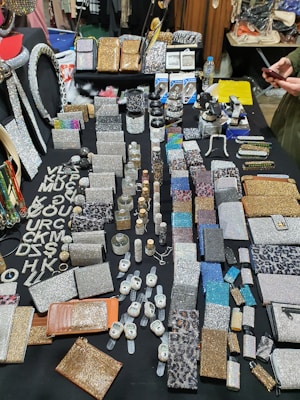 An assortment of sparkling and glittery accessories is displayed on a black table. The items include wallets, vanity cases, compact mirrors, and wristwatches, among others. Many pieces are embellished with rhinestones, sequins, and dazzling patterns. A variety of colors such as silver, gold, and multicolored designs are present, and the overall display is organized in a visually appealing manner. Some text made from shiny material is seen on the left side.