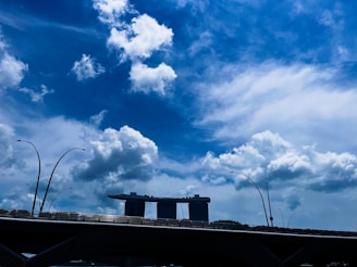 A modern architectural structure with three tall towers connected by a curved platform at the top is seen against a backdrop of a vibrant blue sky with scattered clouds. In the foreground, there is a bridge with several streetlights curving elegantly upwards.