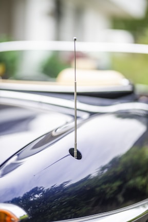 A close-up view of a car's exterior, featuring a sleek metallic antenna. The surface is shiny and reflective, capturing blurred reflections of the surrounding greenery and sky.