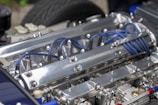 A close-up of a polished metal engine with visible blue ignition wires and various metallic components. The engine features prominent bolts and a sleek, reflective surface with detailed mechanical parts.