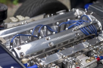 A close-up of a polished metal engine with visible blue ignition wires and various metallic components. The engine features prominent bolts and a sleek, reflective surface with detailed mechanical parts.