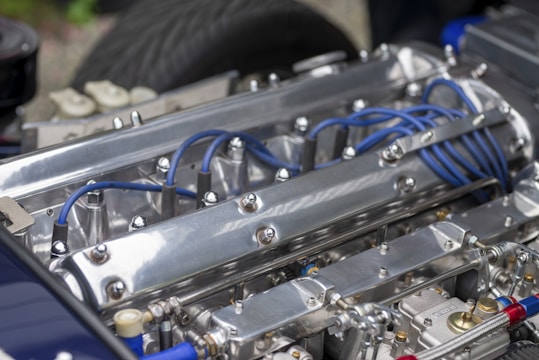 A close-up of a polished metal engine with visible blue ignition wires and various metallic components. The engine features prominent bolts and a sleek, reflective surface with detailed mechanical parts.